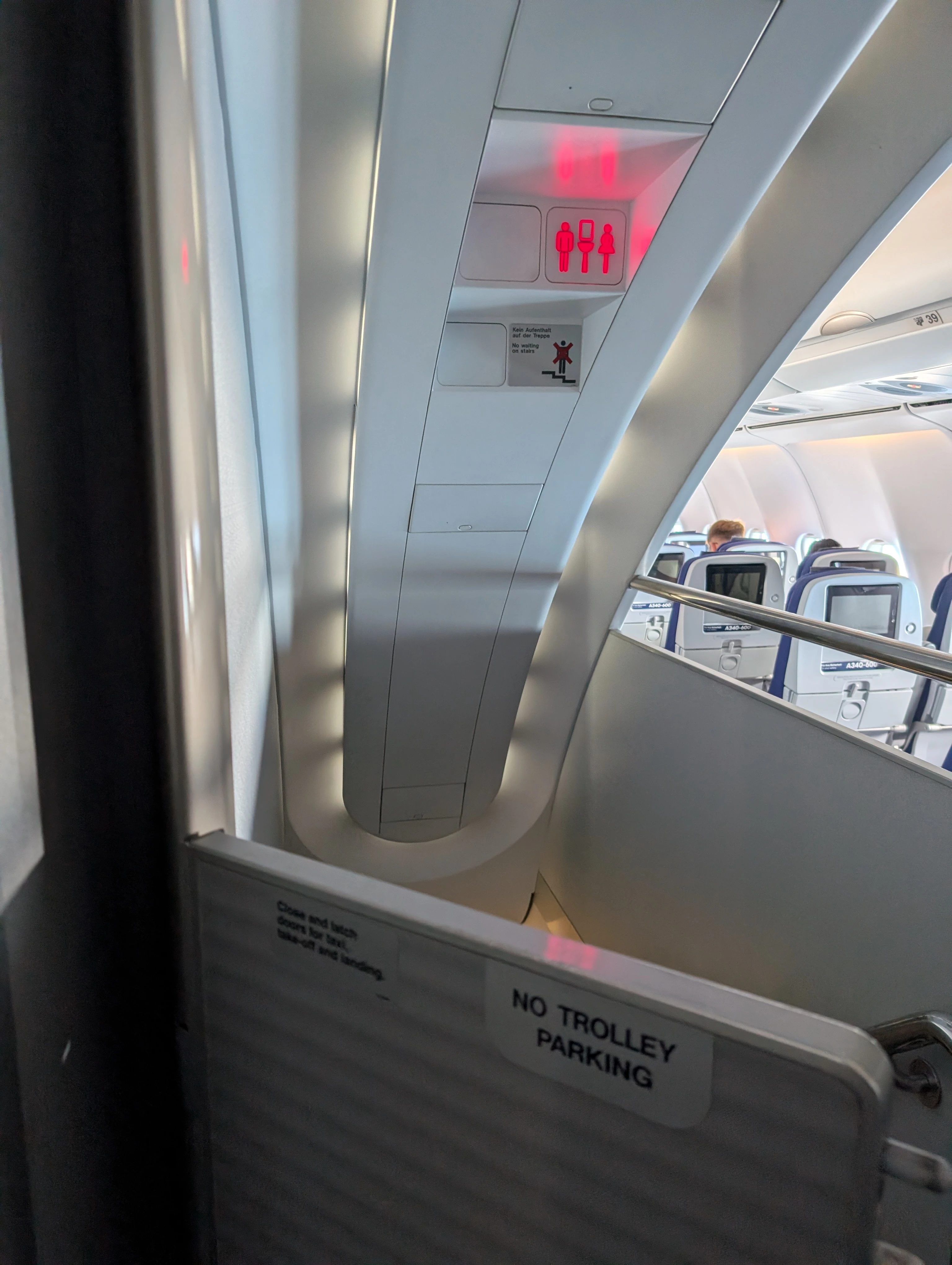 The curved staircase down to the lower-deck lavatories of the A340-600, with a red lavatory sign, a &lsquo;No waiting on stairs&rsquo; notice