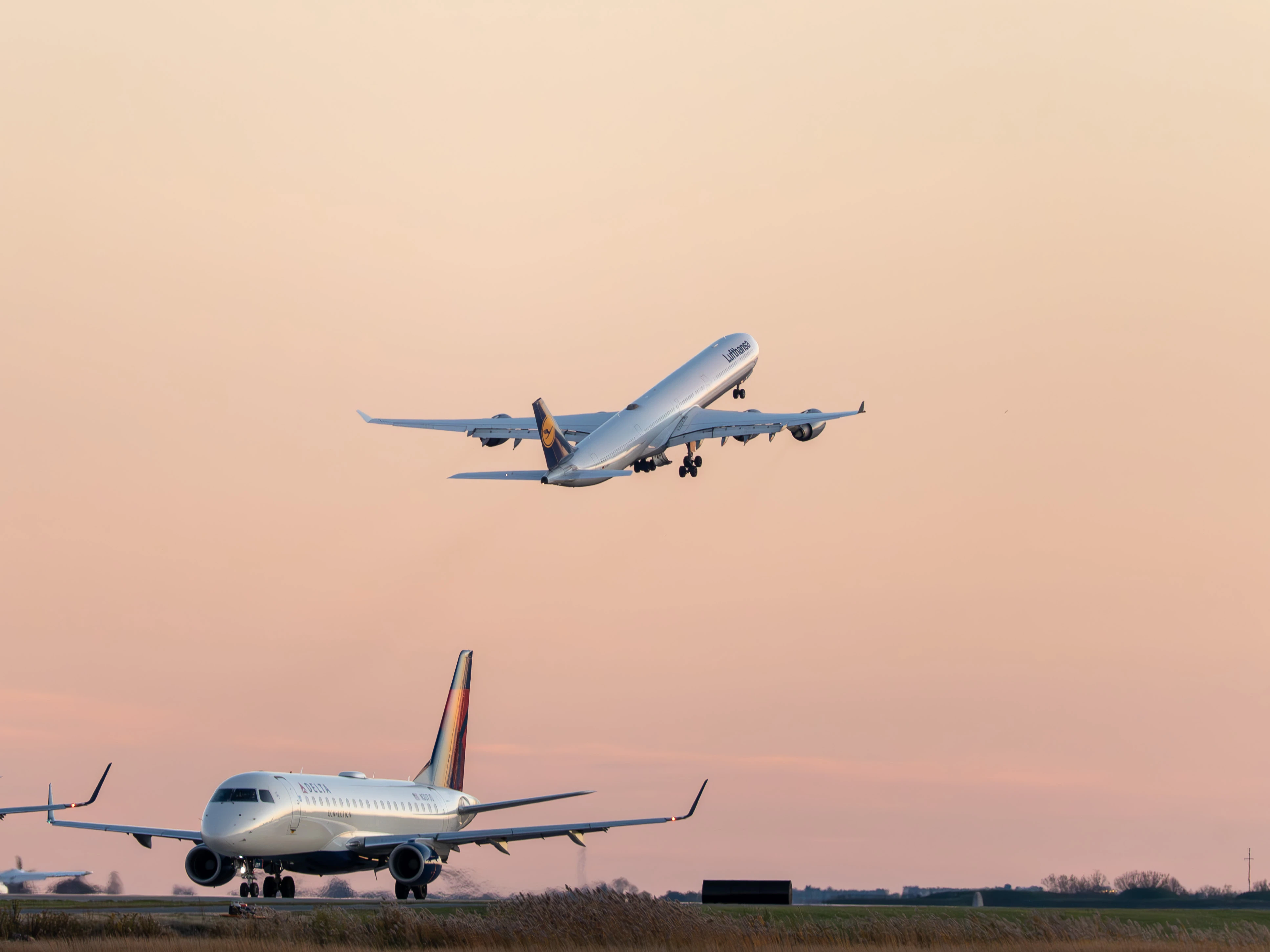 Lufthansa A340-600 climbing steeply into the pink twilight sky while a Delta regional jet waits on the ground